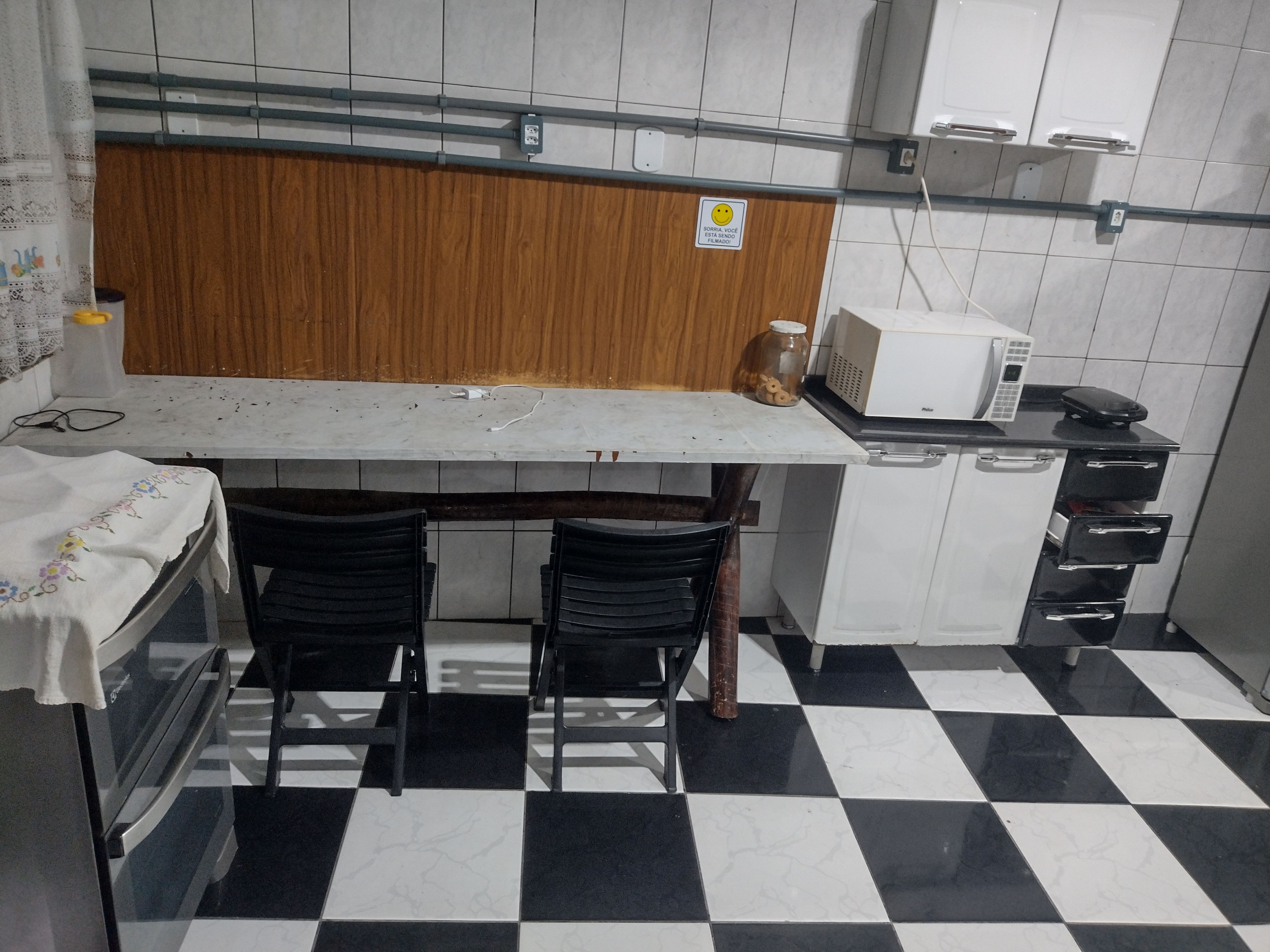 A small kitchenette with a microwave, a coffee maker, and a refrigerator. There are two chairs and a table with a white tablecloth. The floor has a black and white checkered pattern.