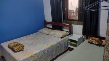 Una habitación pequeña con una cama individual, cabecera de madera, una ventana con cortinas, una mesita de noche y un estante en la pared.