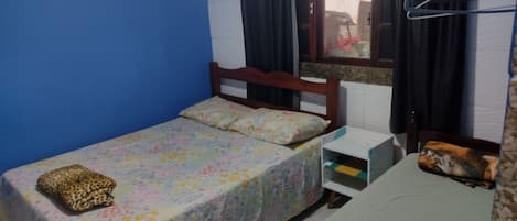A small room with a single bed, a wooden headboard, a window with curtains, a small bedside table, and a wall-mounted shelf.