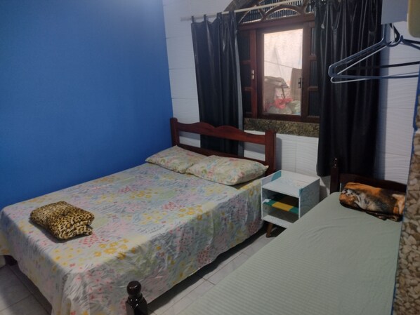 A small room with a single bed, a wooden headboard, a window with curtains, a small bedside table, and a wall-mounted shelf. - Recanto dos Loureiros (Saquarema)