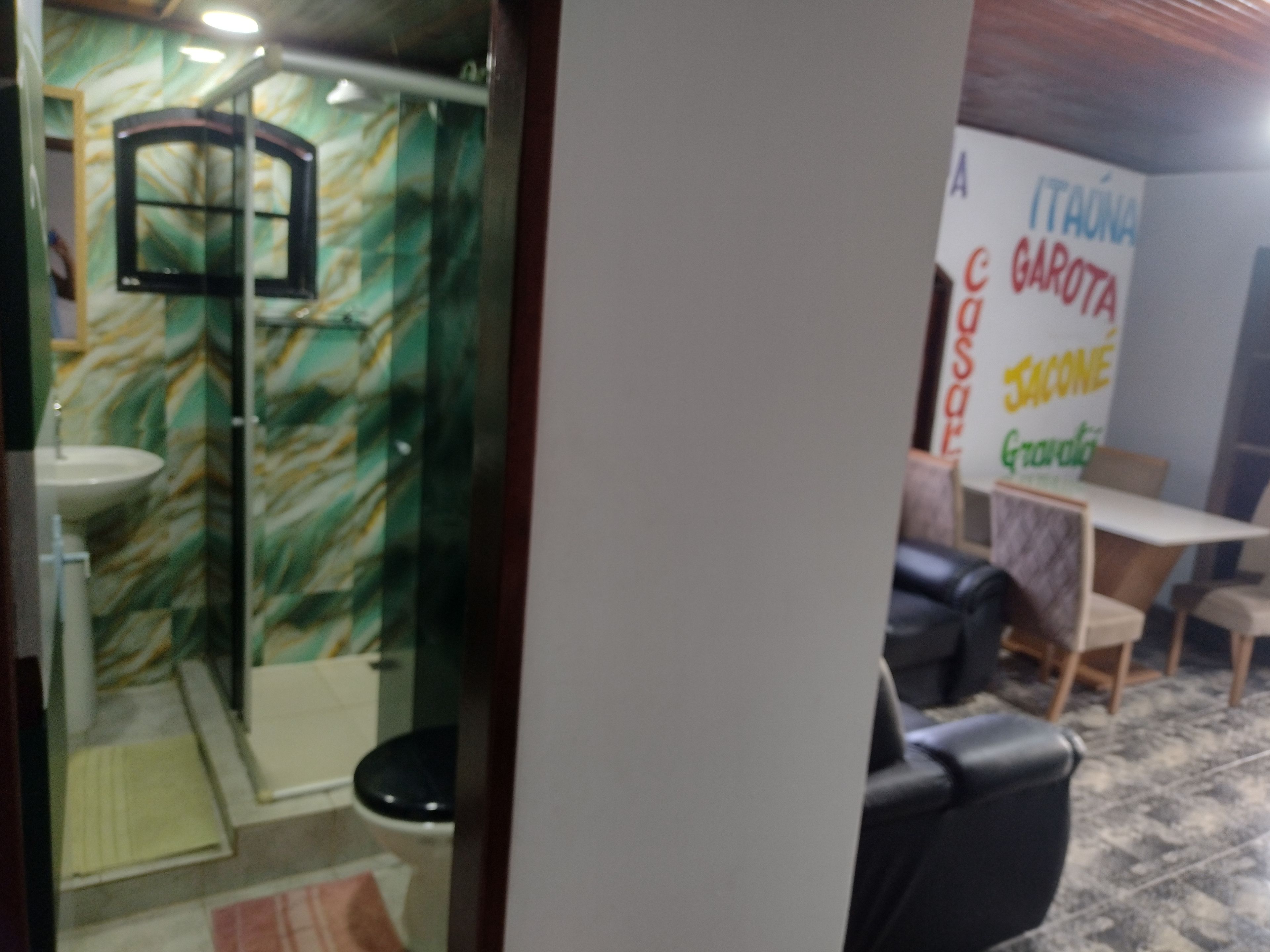 Un baño con lavamanos, espejo y ducha. Se observa una pared con un diseño verde y blanco. En primer plano, hay una mesa y sillas.