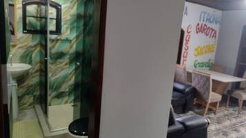 Un baño con lavamanos, espejo y ducha. Se observa una pared con un diseño verde y blanco. En primer plano, hay una mesa y sillas.