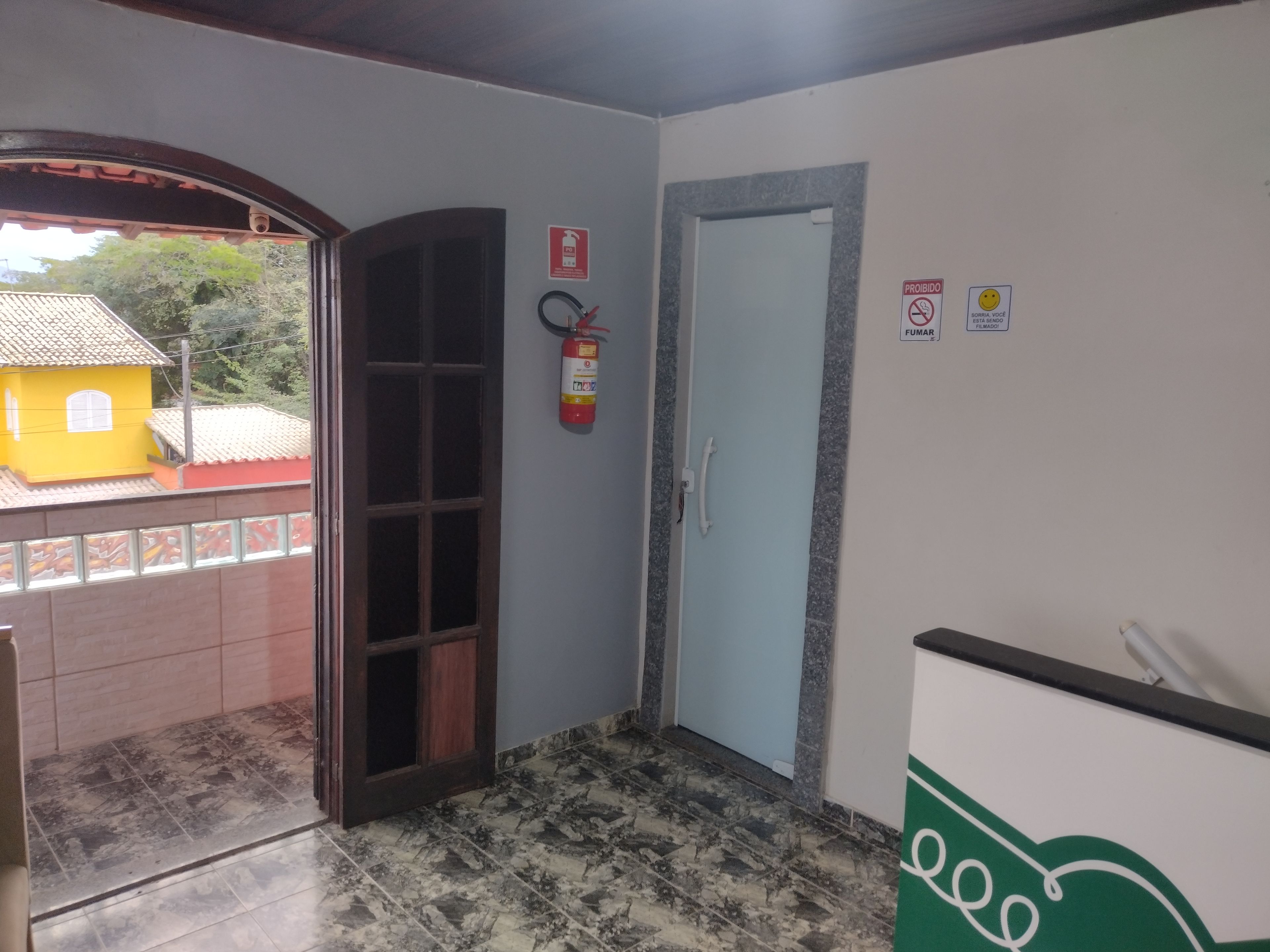 Corredor com extintor, porta e vista para um edifício amarelo lá fora.