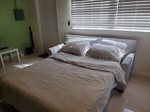 A bedroom with a bed, a bedside table, a power outlet, and a window with blinds. - Remodeled Artist Studio in Coconut Grove (Miami)