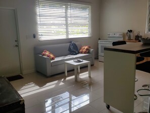 A modern kitchen with a sofa and a small table, a refrigerator, and a window with blinds. - Remodeled Artist Studio in Coconut Grove (Miami)