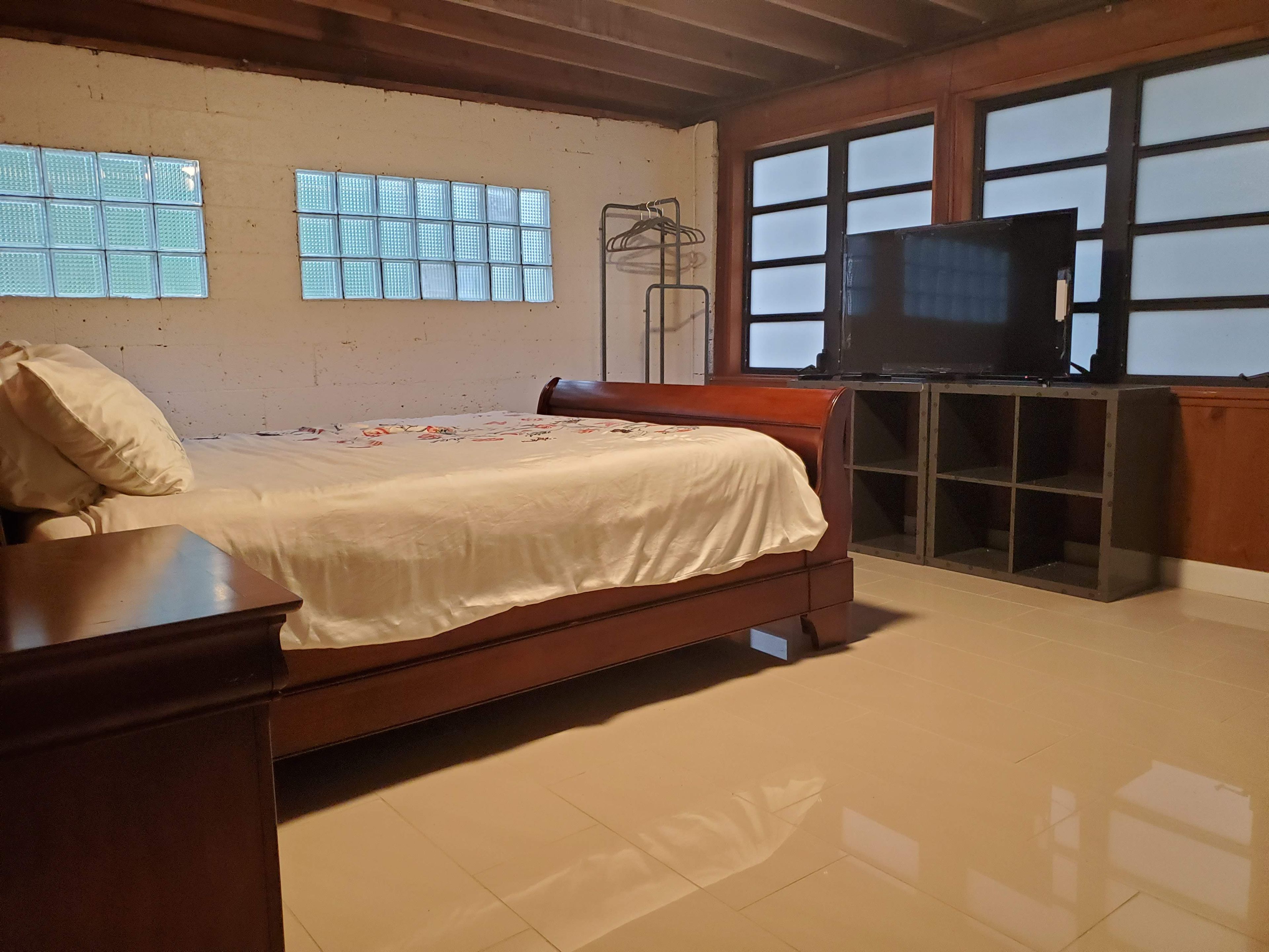 Un dormitorio con una cama, un tocador de madera, un televisor sobre un mueble bajo y una ventana con persianas.