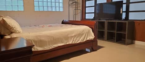Un dormitorio con una cama, un tocador de madera, un televisor sobre un mueble bajo y una ventana con persianas.