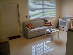Un espace de vie compact comprenant un canapé, une table basse, une cuisinière et une fenêtre avec des stores.