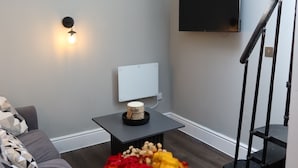 Ein Wohnzimmer mit einem an der Wand befestigten Flachbildfernseher, einem kleinen Tisch mit einer Blumenvase und einer Treppe zur rechten Seite.