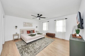 Ein modernes Schlafzimmer mit Bett, Nachttischen, Deckenventilator, Fernseher und einem Fenster mit Vorhängen.