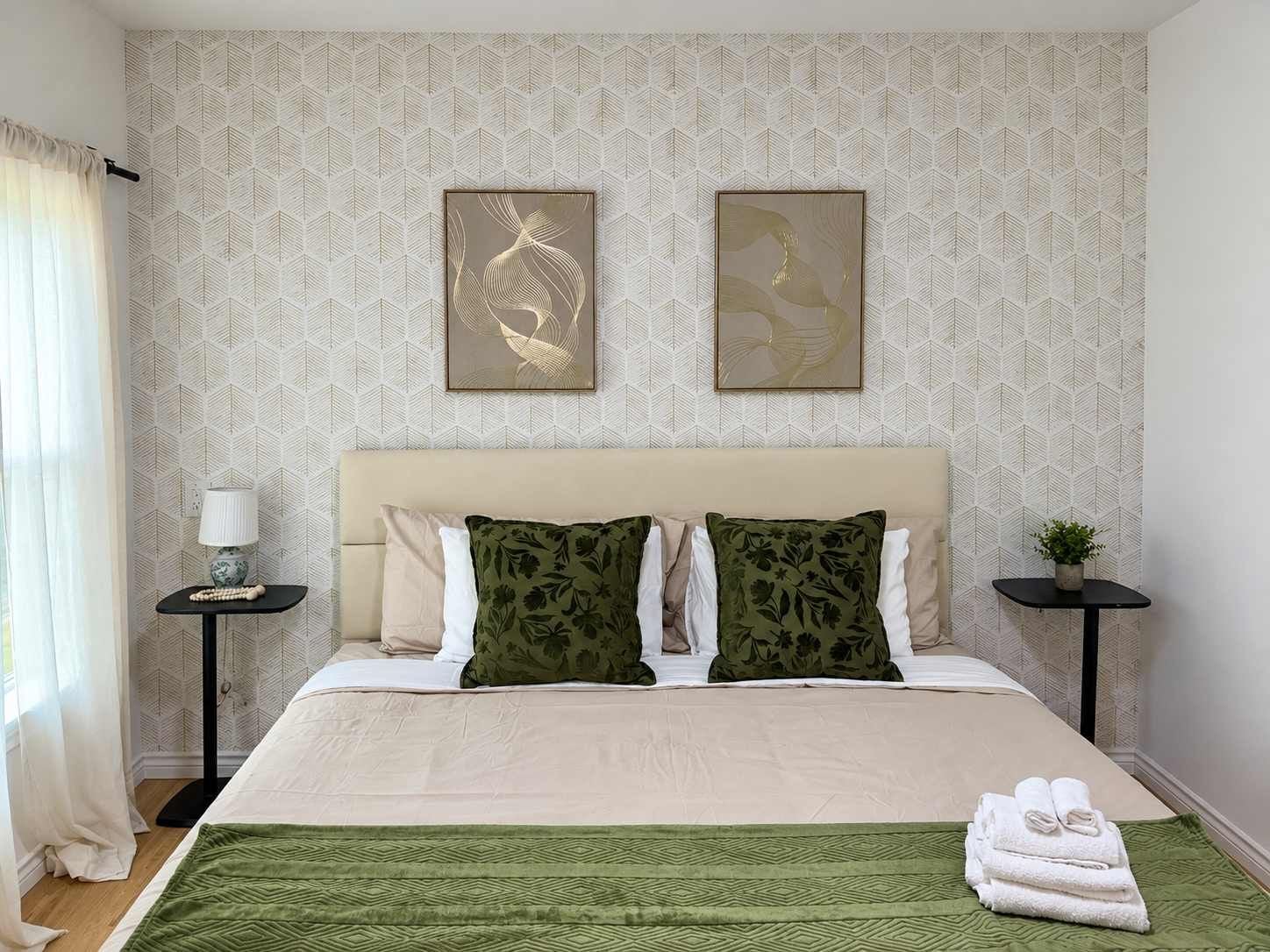 A hotel room with a bed, two pillows, a bedside table with a lamp, a wall with leaf-pattern wallpaper, and two framed artworks.