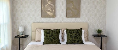 A hotel room with a bed, two pillows, a bedside table with a lamp, a wall with leaf-pattern wallpaper, and two framed artworks.