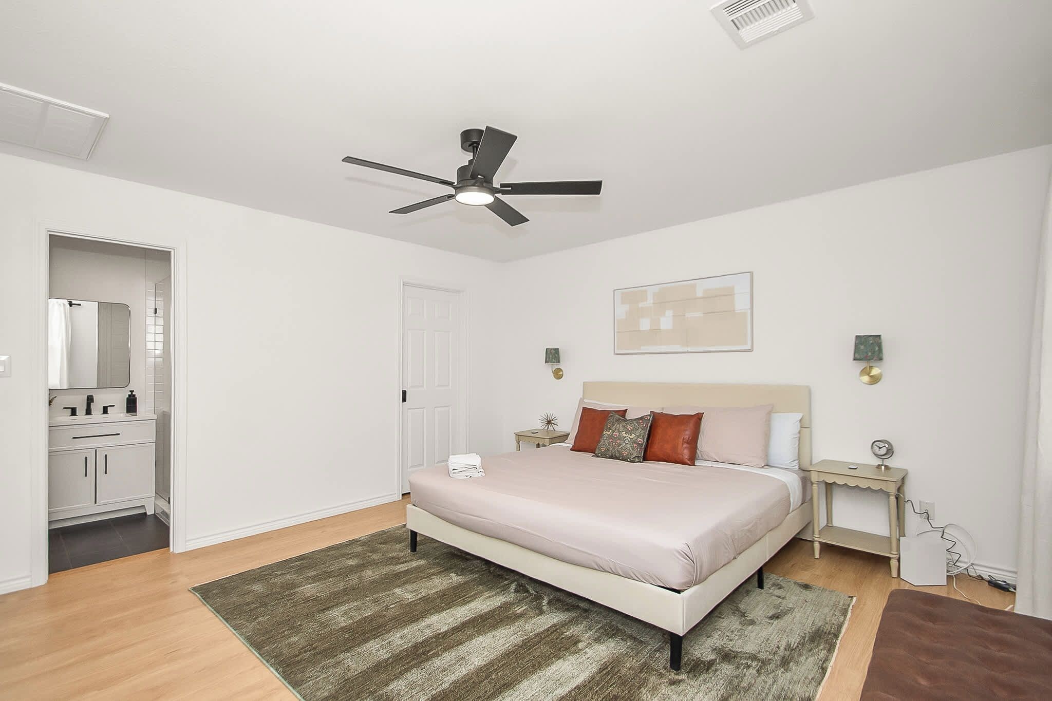 A bedroom with a bed, a ceiling fan, a nightstand, a wall-mounted lamp, and a door leading to a bathroom.