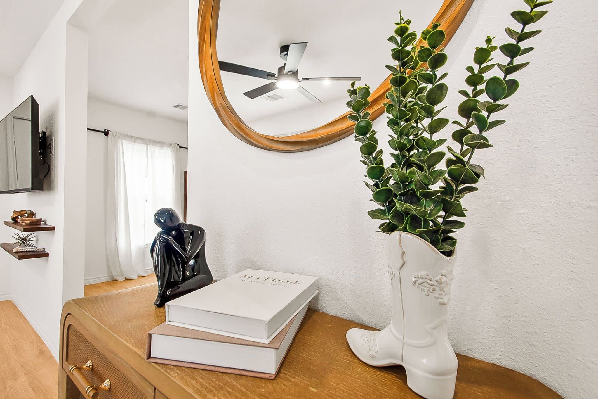 Een kamer met een houten dressoir, een witte vaas met groen bladwerk, een plafondventilator en een spiegel.