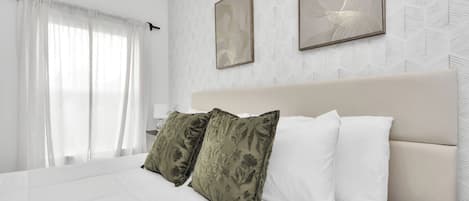 A hotel room with a bed, white curtains, a headboard, and two framed artworks on the wall.