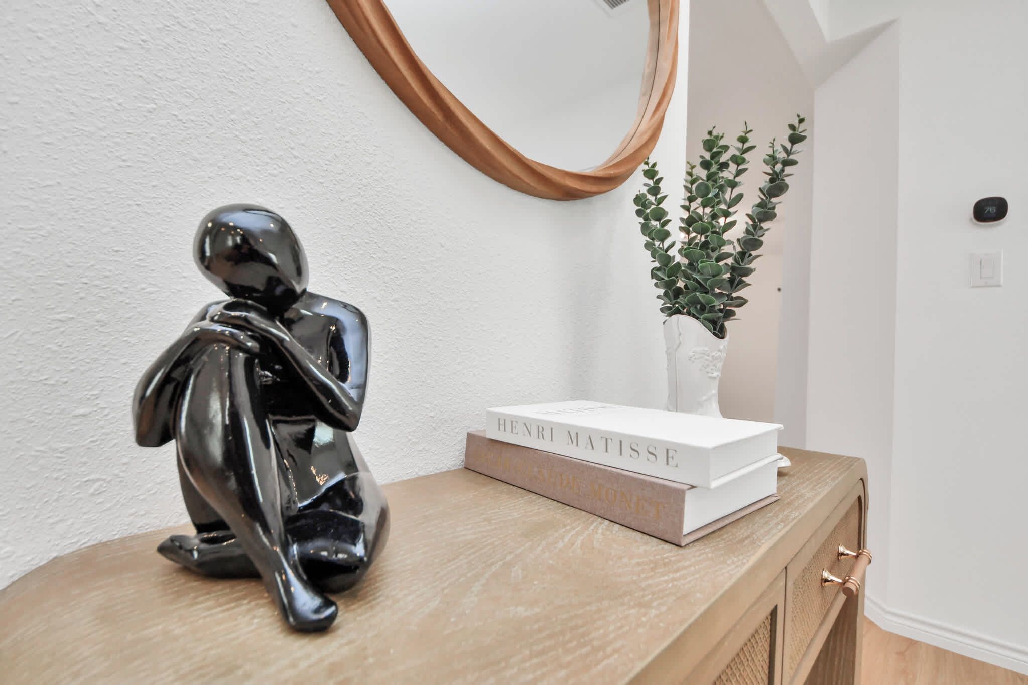 Een strak ingerichte kamer met een houten console, een zwart abstracte sculptuur, een witte vaas met groene planten en een spiegel.