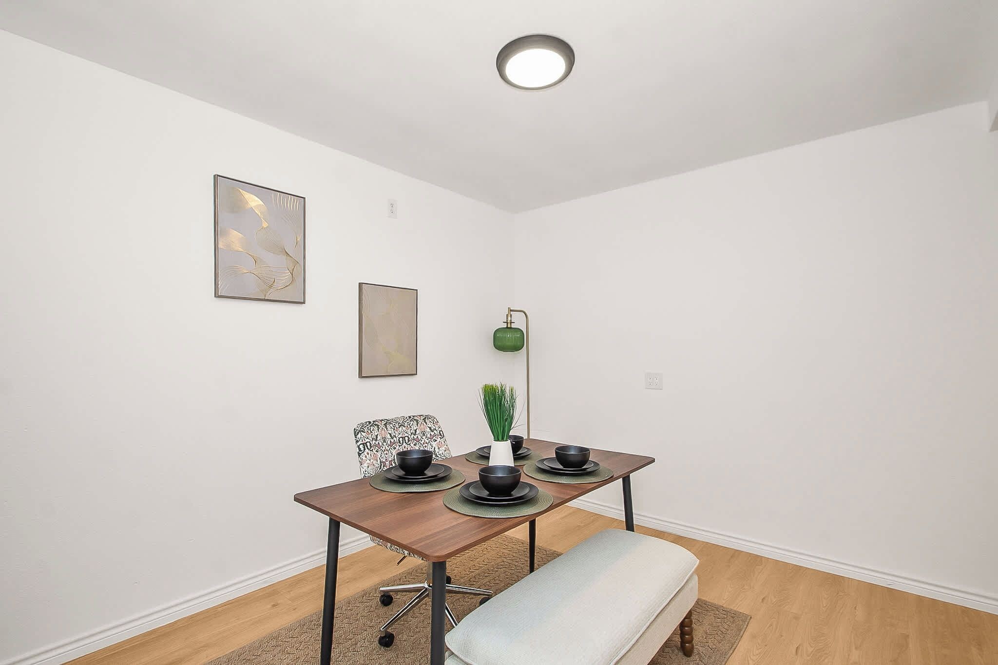 A dining area with a table set for four, a chair, a bench, a floor lamp, and two wall-mounted artworks.