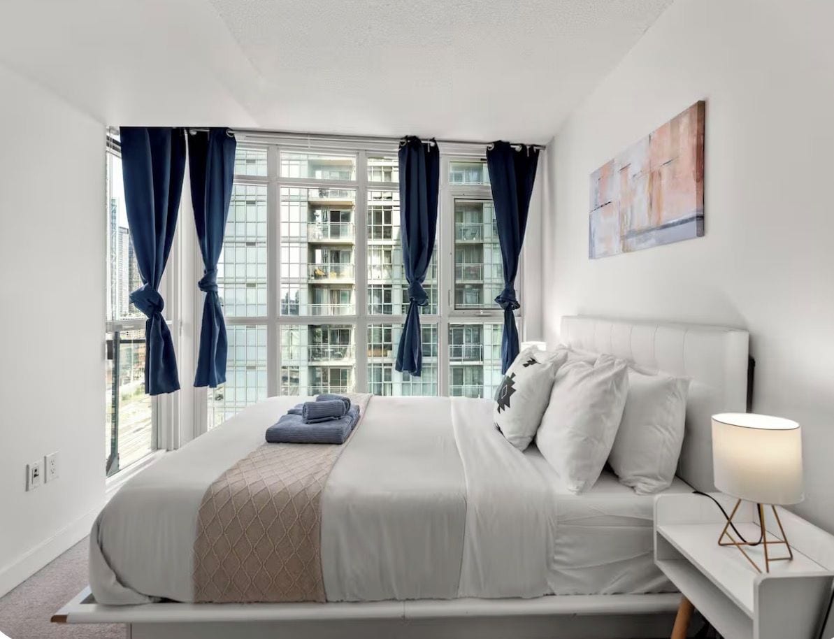 A modern bedroom with a large bed, a bedside table with a lamp, and a view of buildings through the window.