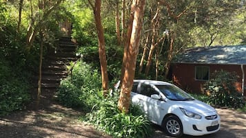 Un coche blanco estacionado cerca de un edificio rodeado de abundante follaje y una escalera que sube.