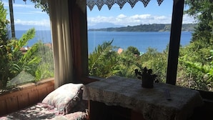 A room with a bed, a table with a lace tablecloth, and a view of a lake and mountains. - Cabañas el Bosque Patagónico (Frutillar)