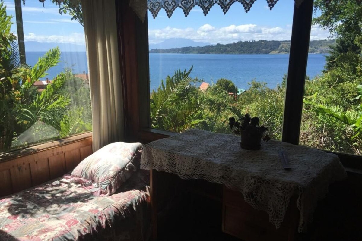 Une chambre avec un lit, une table recouverte d’une nappe en dentelle, et une vue sur un lac et des montagnes.