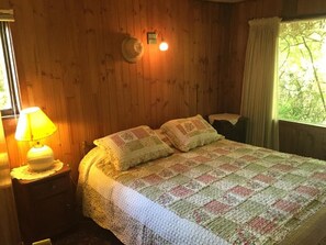 Una habitación de madera con una cama, una mesita de noche con lámpara y una ventana con cortinas.