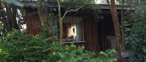 A rustic wooden cabin with a porch, surrounded by lush greenery and flowering plants.