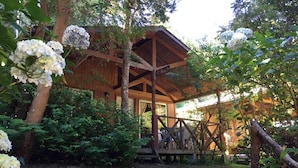 A wooden cabin with a covered porch, surrounded by lush greenery and white flowers. - Cabañas el Bosque Patagónico (Frutillar)