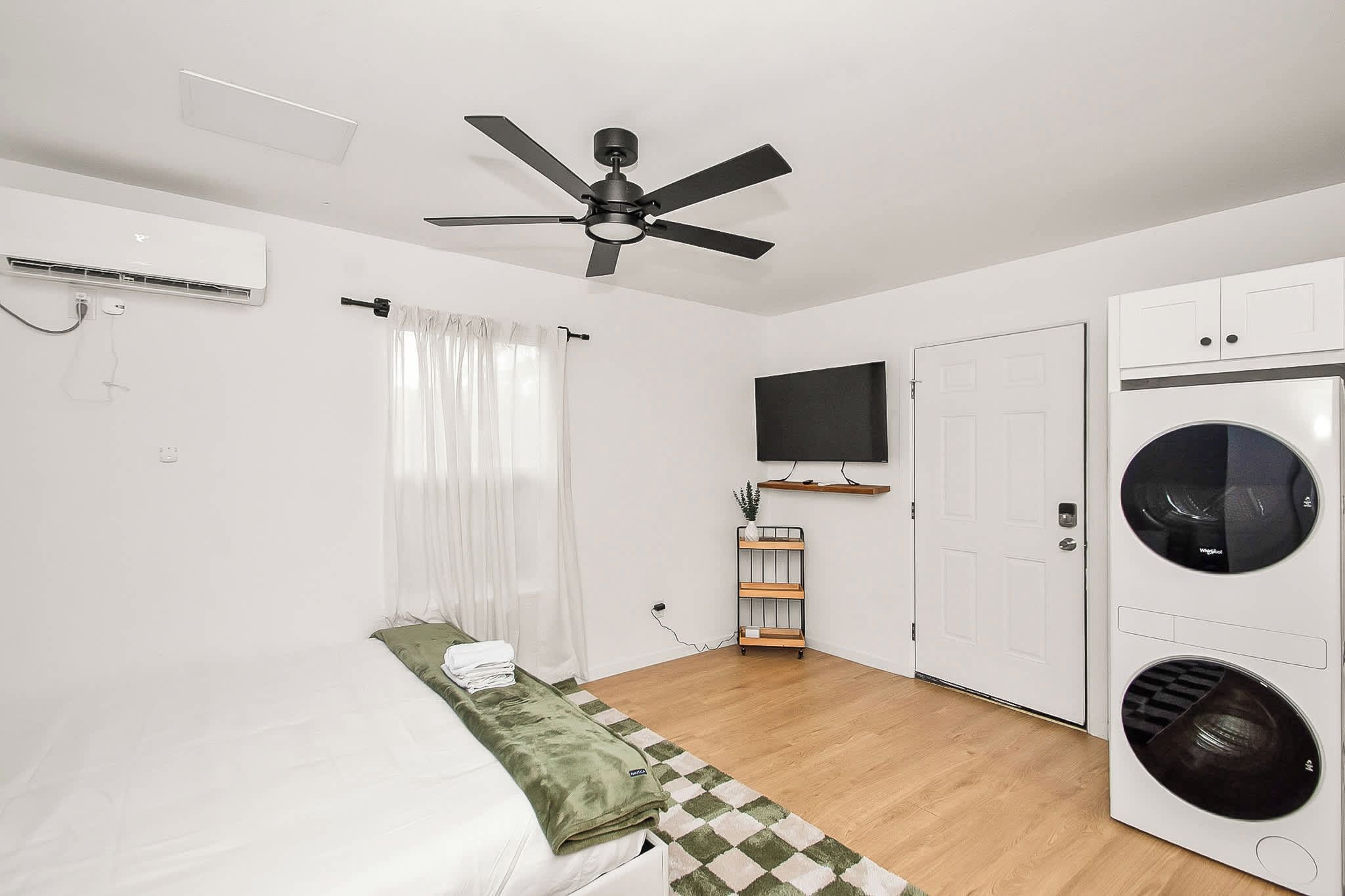A bedroom with a bed, a ceiling fan, a TV, a washing machine, and a door.