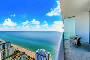 Ein Balkon mit Blick auf den Ozean und den Strand, ein Essbereich mit Korbmöbeln und ein modernes Gebäude mit Glasfassade.