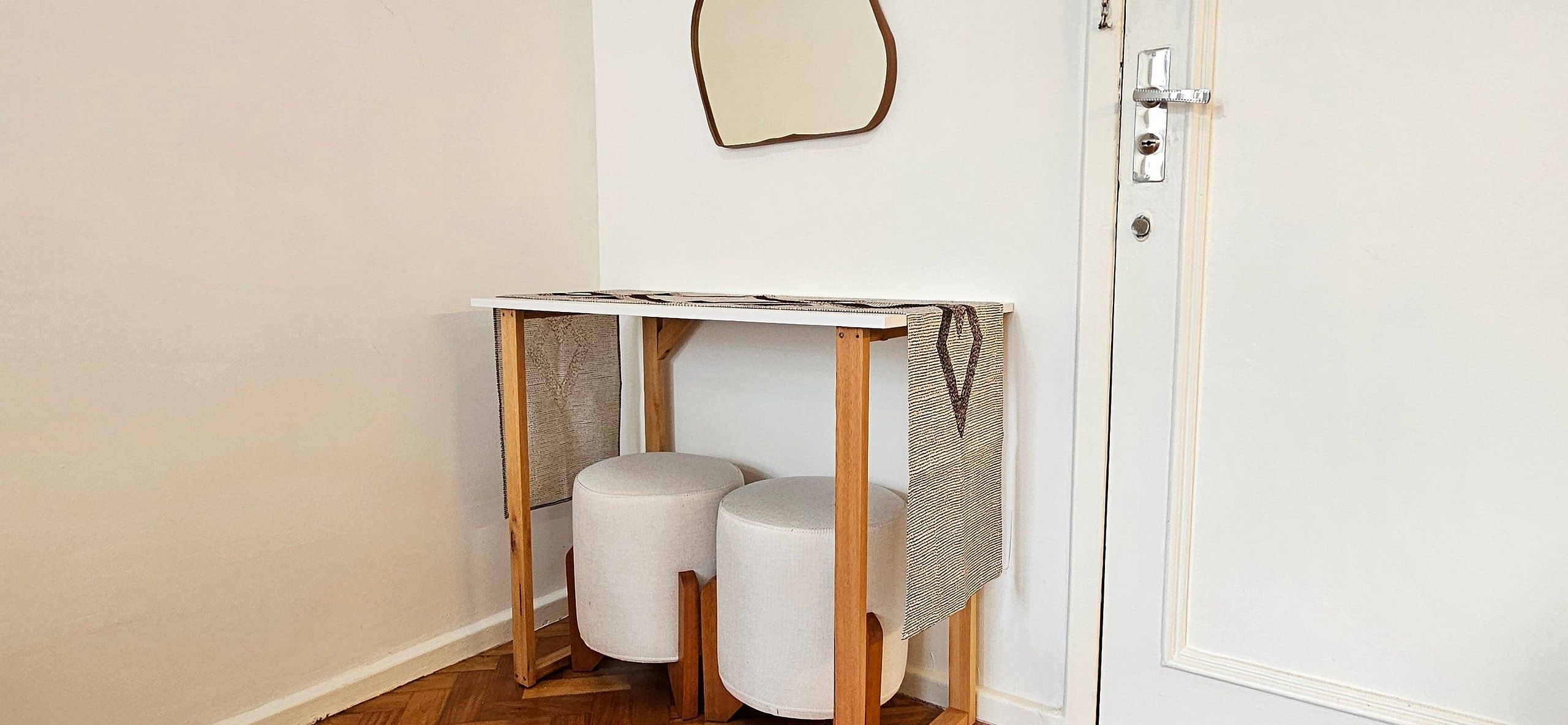 Uma mesa pequena de madeira branca com tampo estampado, dois banquinhos cilíndricos brancos e uma porta branca simples com puxador prateado.