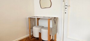 Una mesa pequeña de madera blanca con un tablero estampado, dos taburetes blancos cilíndricos, y una puerta blanca sencilla con un picaporte plateado.
