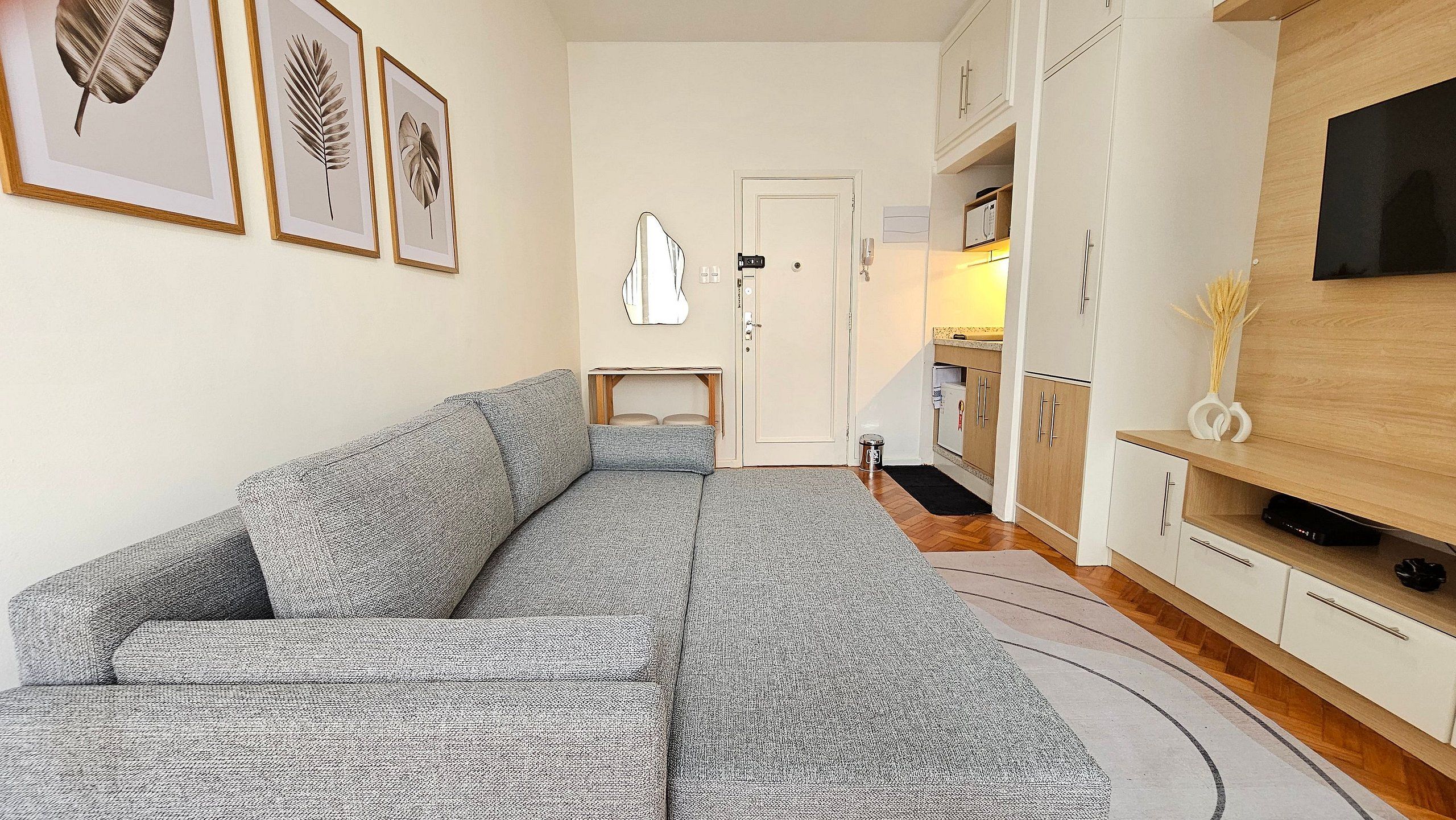 Una sala moderna con un sofá gris estampado, un televisor de pantalla plana montado en la pared y una cocina con gabinetes de madera y encimera.