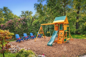 A playground with a swing set and slide, surrounded by trees and a wooden deck.