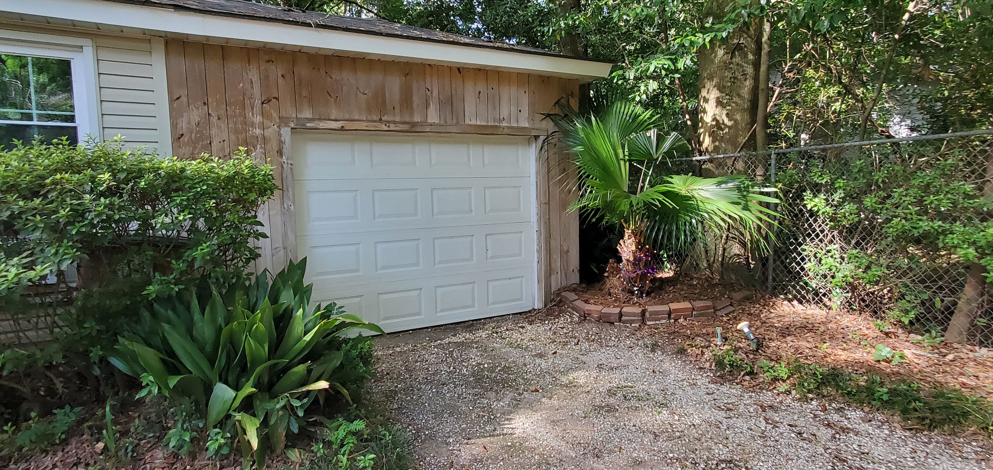 A garage with a white door, surrounded by greenery and a gravel driveway.