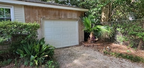 Un garage avec une porte blanche, entouré de verdure et bordé d’une allée en gravier.