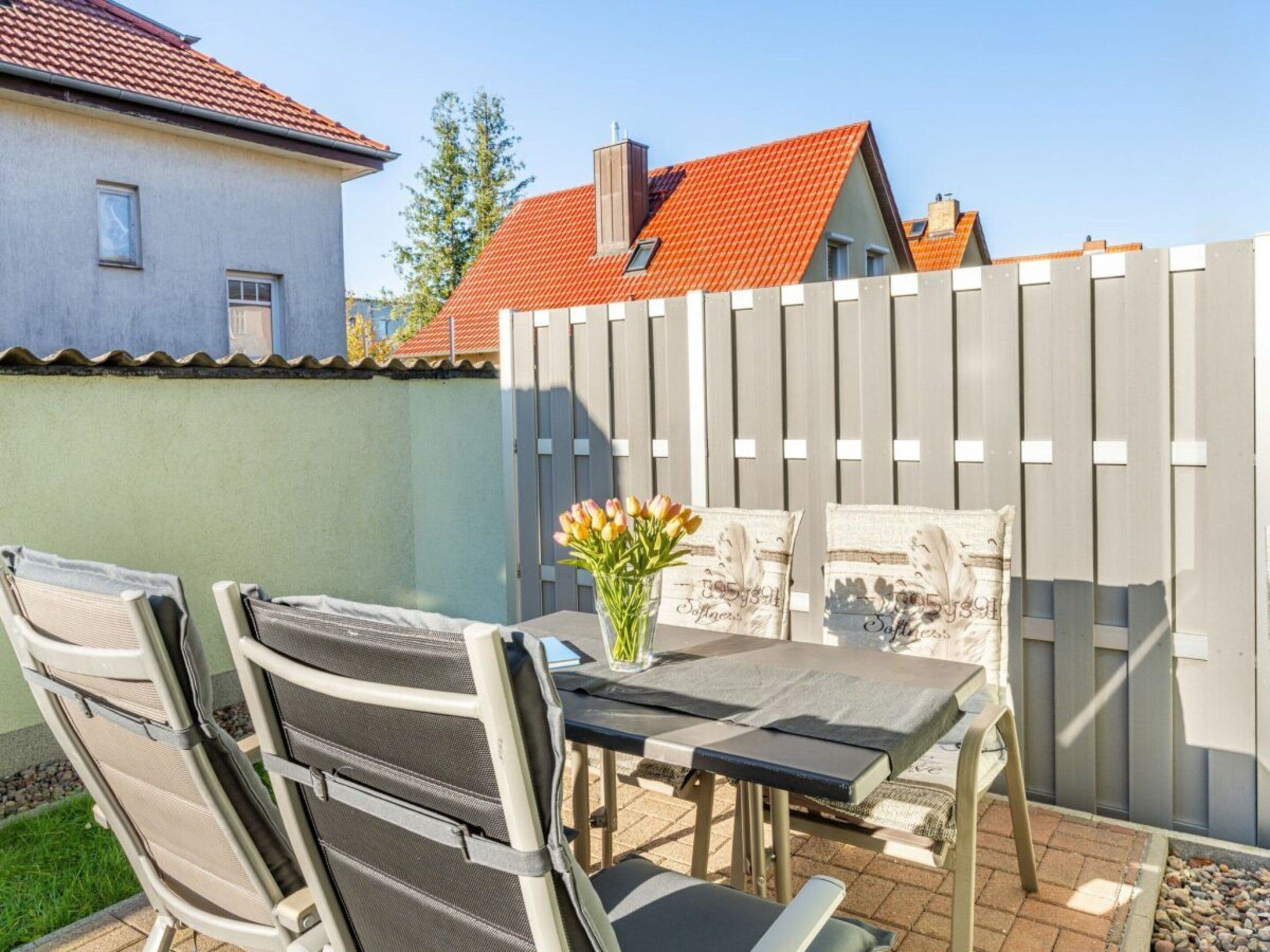 An outdoor patio with a table and chairs, a vase with flowers, and a wooden fence.