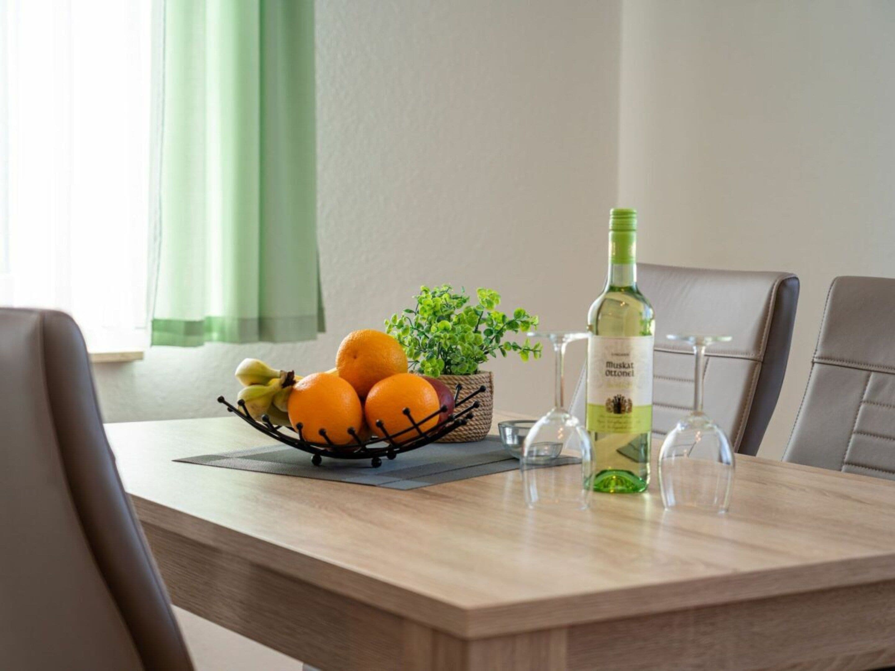 A dining table with a bottle of Muskat Ottonel wine, a fruit basket with oranges and bananas, and a small potted plant.
