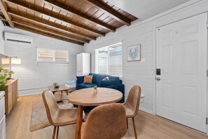 Una habitación con techo de madera, paredes de ladrillo blanco, un sofá azul, una mesa redonda de madera y dos sillas.
