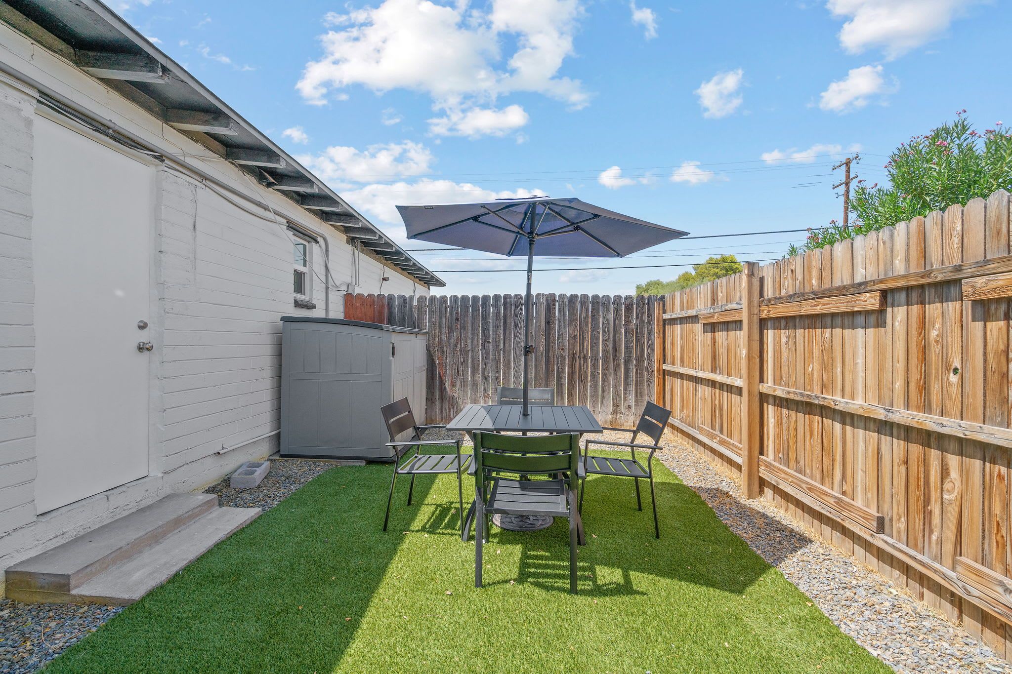 A backyard with a table and chairs, a patio umbrella, a wooden fence, and a white building.