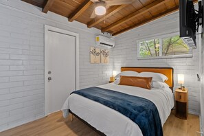 Un dormitorio con techo de madera, paredes de ladrillo blanco, cama con ropa de cama blanca y azul, dos lámparas de pared y una ventana con vistas a árboles.