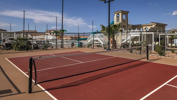 En udendørs tennisbane med net, omgivet af et hegn, og en bygning med et tårn i baggrunden.