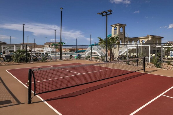 An outdoor tennis court with a net, surrounded by a fence, and a building with a tower in the background.