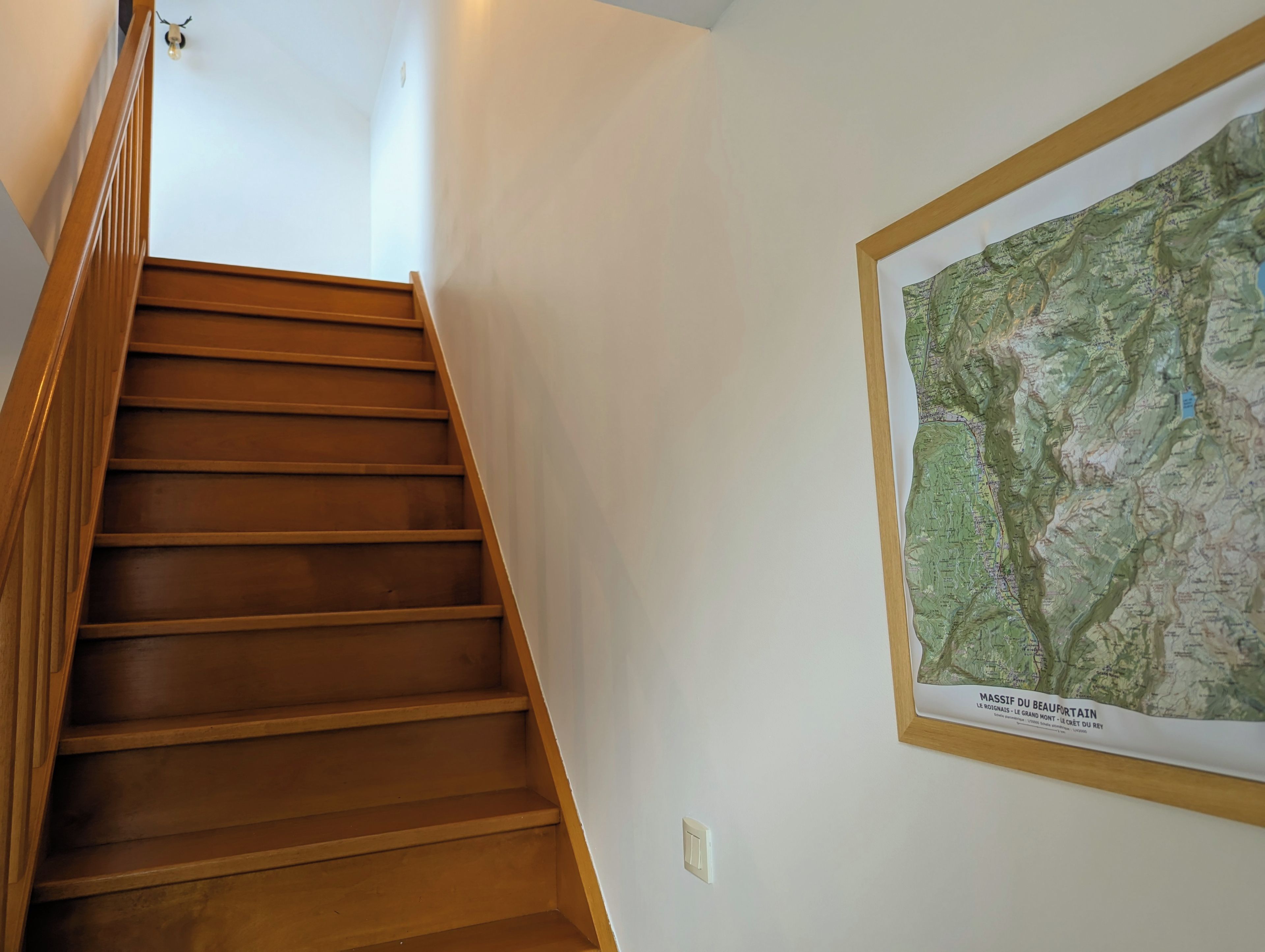 Un escalier en bois menant à l’étage supérieur, un mur blanc et une carte encadrée accrochée au mur.