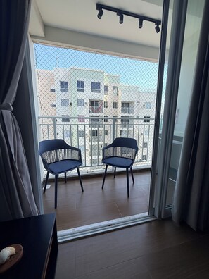 Ein Balkon mit zwei Stühlen, Blick auf ein Gebäude mit Balkonen und ein Vorhang, der das Fenster teilweise verdeckt.