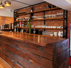 A bar area with a wooden counter, shelves with glasses and bottles, and a brick wall.