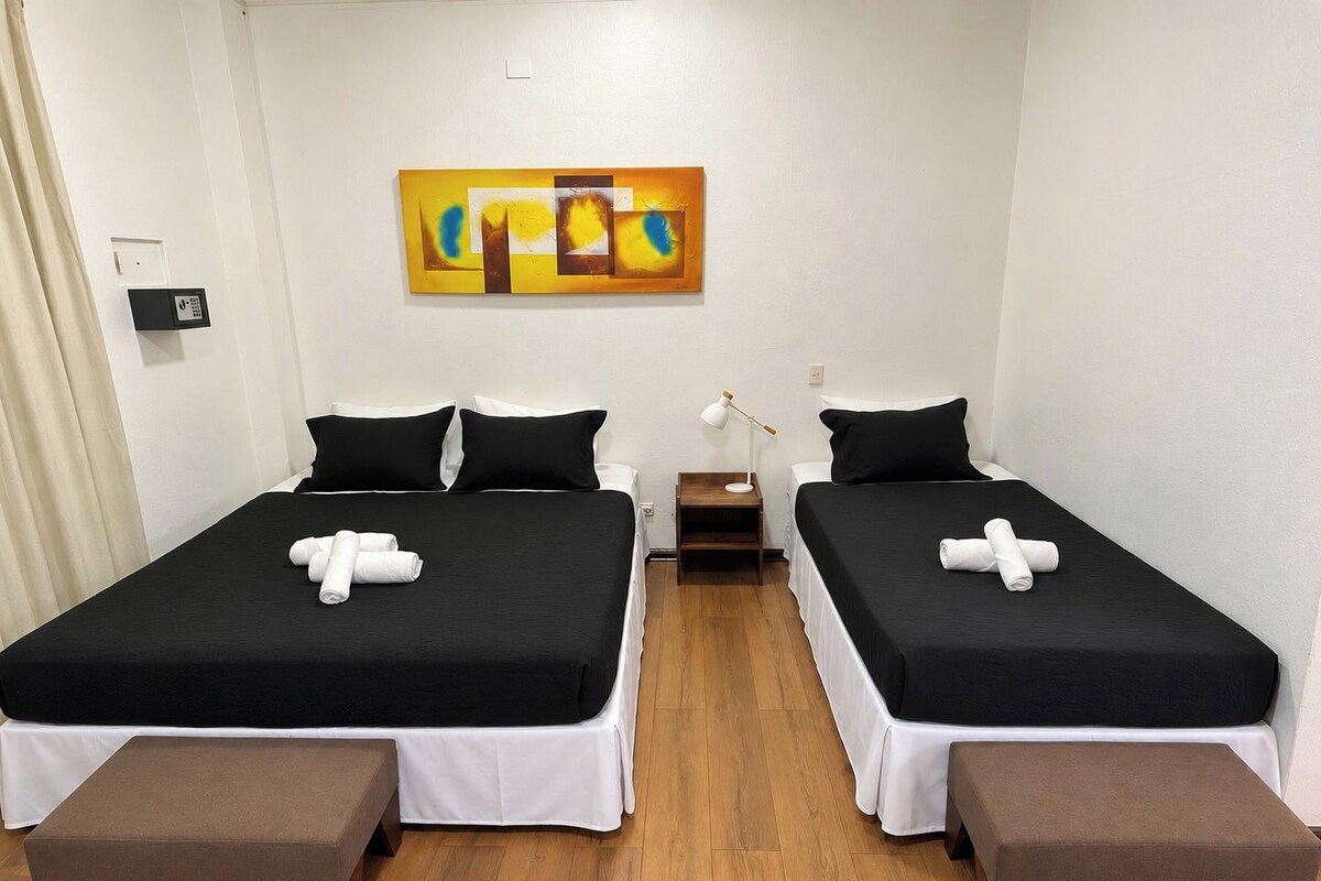 A hotel room with two beds, a painting on the wall, a bedside table with a lamp, and a wooden bench.