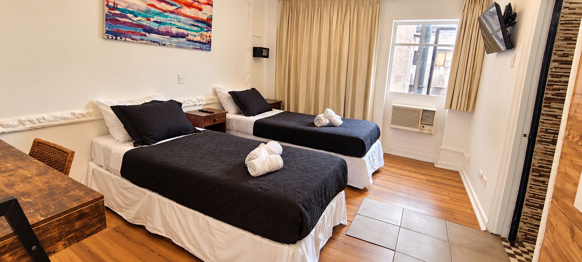 A hotel room with two beds, a wooden table, a painting on the wall, a window with curtains, and a TV mounted on the wall.