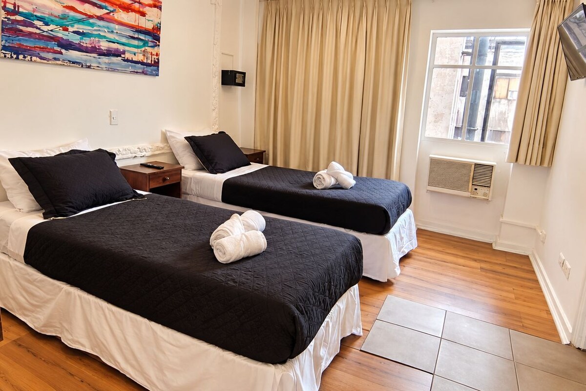 A hotel room with two beds, a wooden table, a painting on the wall, a window with curtains, and a TV mounted on the wall.