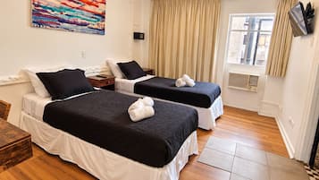 A hotel room with two beds, a wooden table, a painting on the wall, a window with curtains, and a TV mounted on the wall.